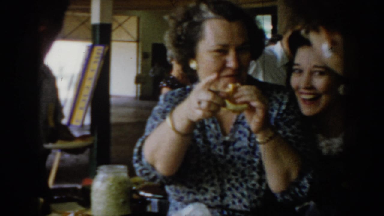 Two women sharing a laugh at a social gathering, circa 1965 Saint Louis; woman in patterned dress holds a sandwich in 8mm color home-movie footage.