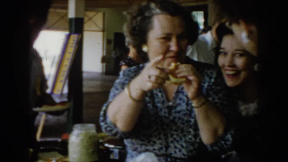 Two women sharing a laugh at a social gathering, circa 1965 Saint Louis; woman in patterned dress holds a sandwich in 8mm color home-movie footage.