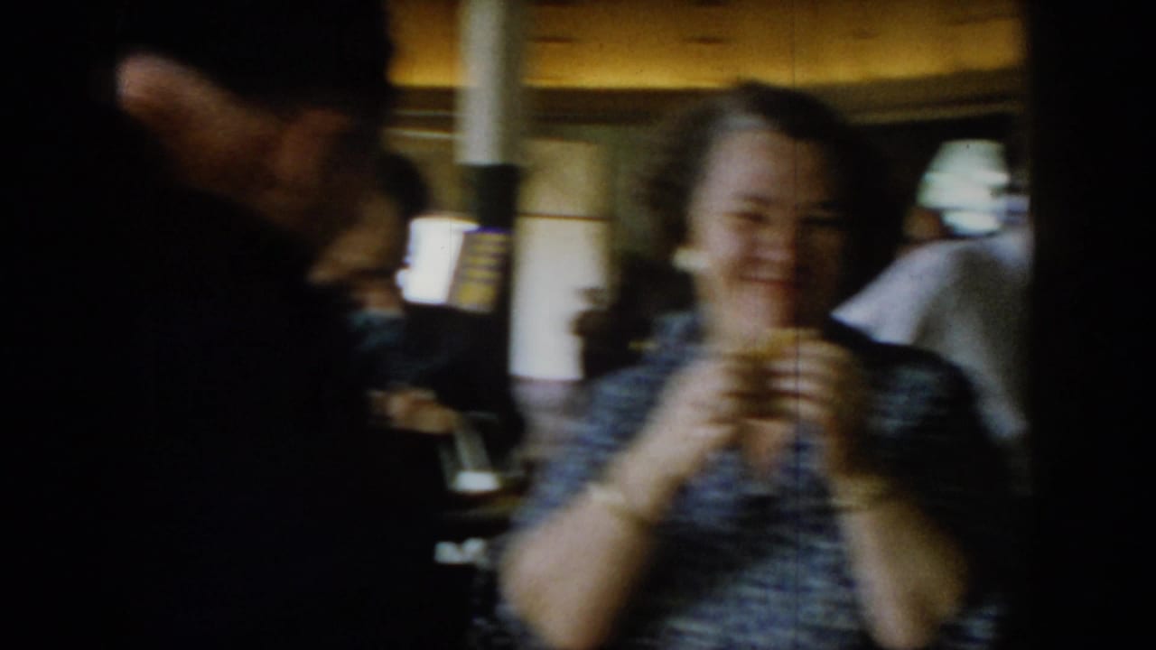 Motion-blurred 8mm frame of a woman laughing and holding food at a social gathering, circa 1965; strong handheld camera movement throughout.