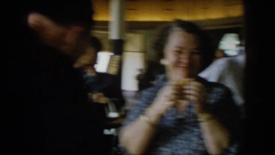 Motion-blurred 8mm frame of a woman laughing and holding food at a social gathering, circa 1965; strong handheld camera movement throughout.