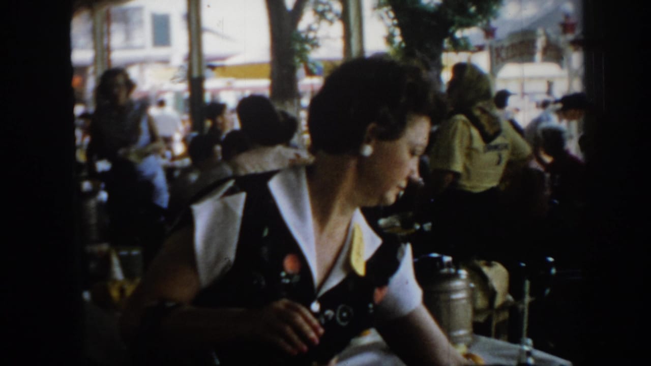 Wider 8mm shot of a woman in a dark jacket at a table in what appears to be an outdoor or semi-outdoor event venue, circa 1965 Saint Louis; crowd and trees behind.