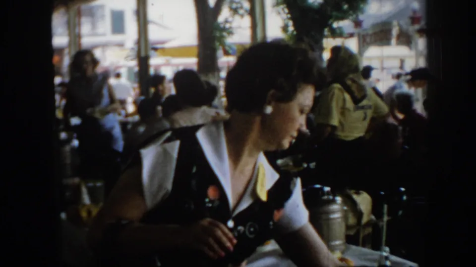Wider 8mm shot of a woman in a dark jacket at a table in what appears to be an outdoor or semi-outdoor event venue, circa 1965 Saint Louis; crowd and trees behind.