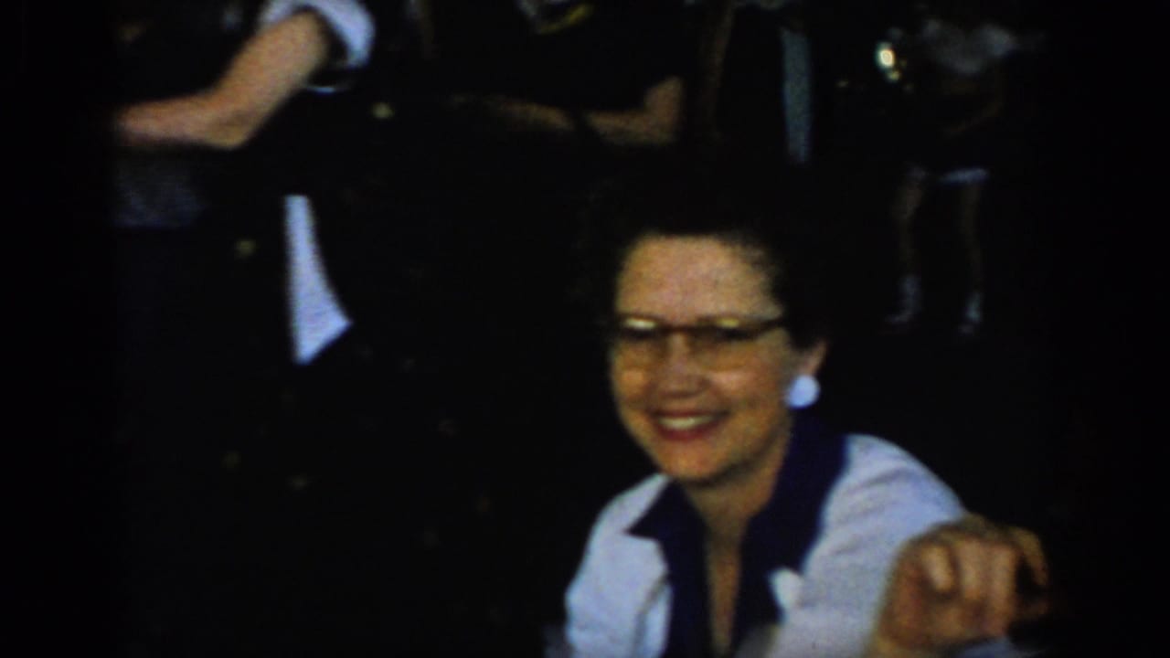 Mid-1960s 8mm home movie: dark-haired woman in cat-eye glasses and white shirt with dark neck-scarf smiles warmly at camera, dim indoor social gathering, St. Louis area, 1965.