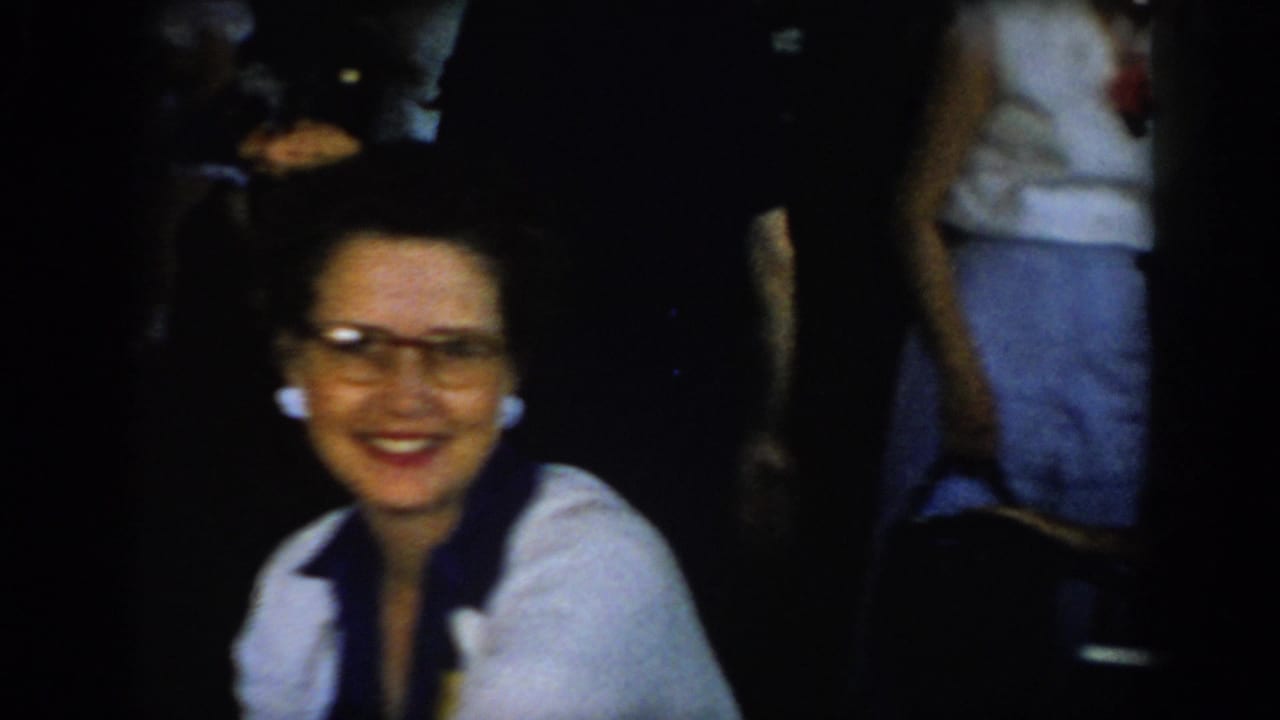 Dark-haired woman in cat-eye glasses and white shirt with dark scarf smiles toward camera; partial figure in white shirt visible at right.