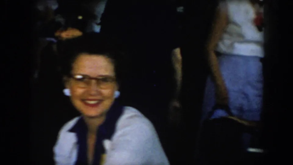 8mm home movie frame: dark-haired woman in cat-eye glasses and white shirt with dark neck-scarf at dim indoor gathering; partial background figure visible at right, St. Louis area, 1965.
