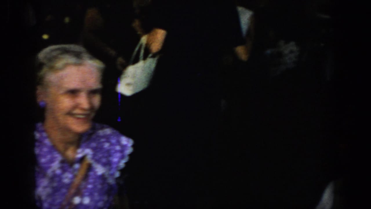8mm home movie frame: older woman with white hair wearing a purple floral print dress and clip-on earrings, carrying a white handbag, at a dim indoor gathering, circa 1965.
