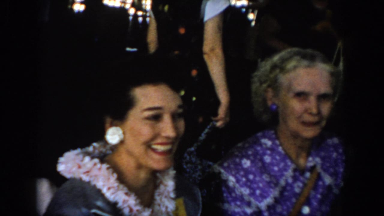 8mm home movie: dark-haired woman in white ruffled blouse with large clip-on earrings smiles beside older woman in purple floral dress; bokeh background lights suggest festive indoor setting, circa 1965.