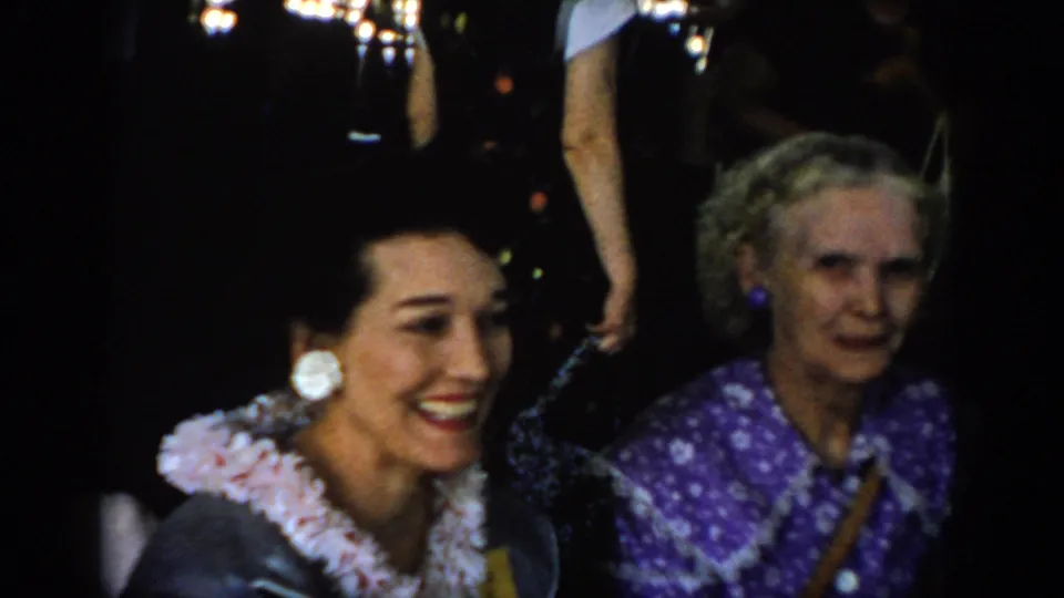8mm home movie: dark-haired woman in white ruffled blouse with large clip-on earrings smiles beside older woman in purple floral dress; bokeh background lights suggest festive indoor setting, circa 1965.