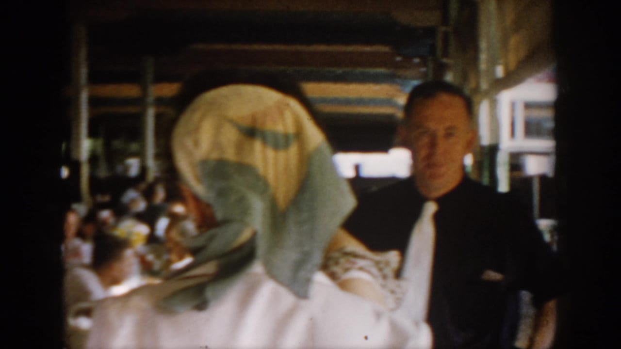 A man in a dark suit and white tie stands in a busy 1965 restaurant dining room in Saint Louis, Missouri, with a woman in white and seated patrons visible around him.