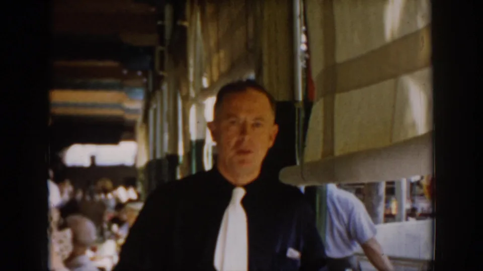 The suited man faces the camera in a mid-1960s restaurant interior in Saint Louis, Missouri; a man in a light blue shirt and multiple seated patrons are visible behind him.