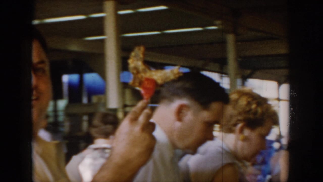 Man in white shirt holds up what appears to be a lollipop or candy at a covered indoor market, circa 1965; concrete pillars, fluorescent strip lights, and blue containers visible in background.