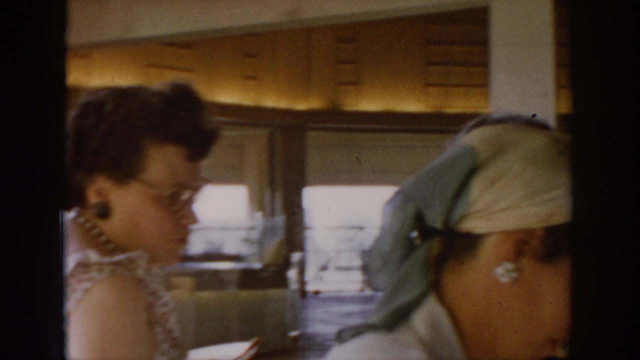 Woman with glasses leans over a market counter at a covered indoor market, Saint Louis, circa 1965; shelved goods and diffuse window light visible in background.