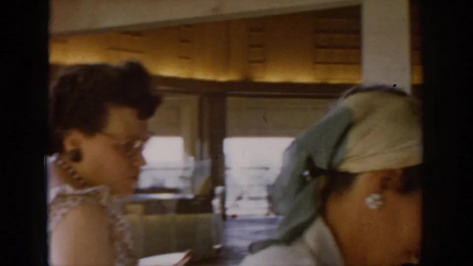 Woman with glasses leans over a market counter at a covered indoor market, Saint Louis, circa 1965; shelved goods and diffuse window light visible in background.