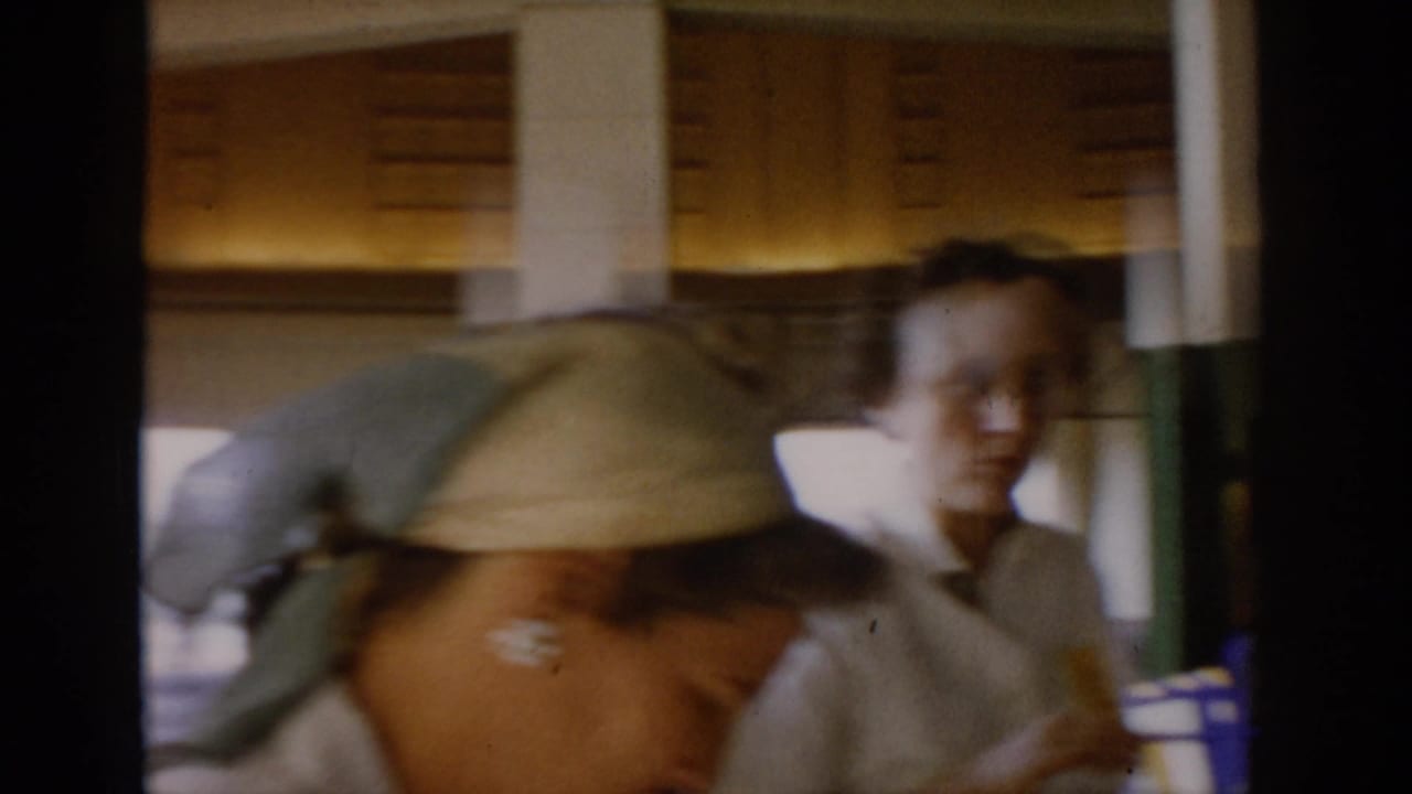 Two women in close-up; one wearing a light headscarf, the other in a pale top, in a warmly lit interior.