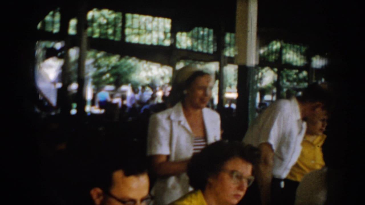 Wide view of a 1960s semi-open pavilion restaurant; woman in white jacket, multiple seated diners, large open windows onto summer trees, Saint Louis circa 1965.