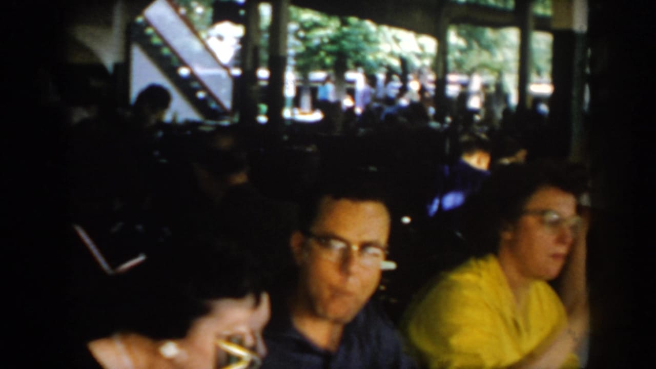 Man with glasses in dark jacket and woman in yellow at a 1960s pavilion dining table; open windows onto summer greenery, Saint Louis circa 1965.
