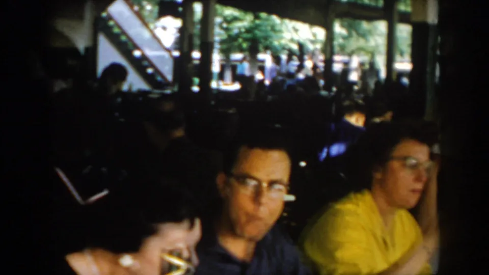 Man with glasses in dark jacket and woman in yellow at a 1960s pavilion dining table; open windows onto summer greenery, Saint Louis circa 1965.