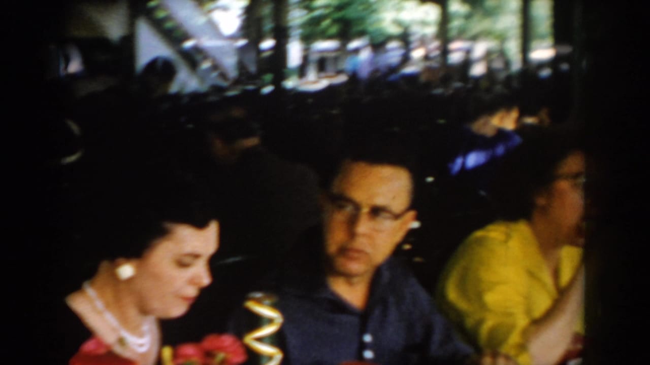 Group of adults seated at long tables in a covered patio restaurant; woman in pearl necklace at left, man with cat-eye glasses at center, woman in yellow blouse behind.