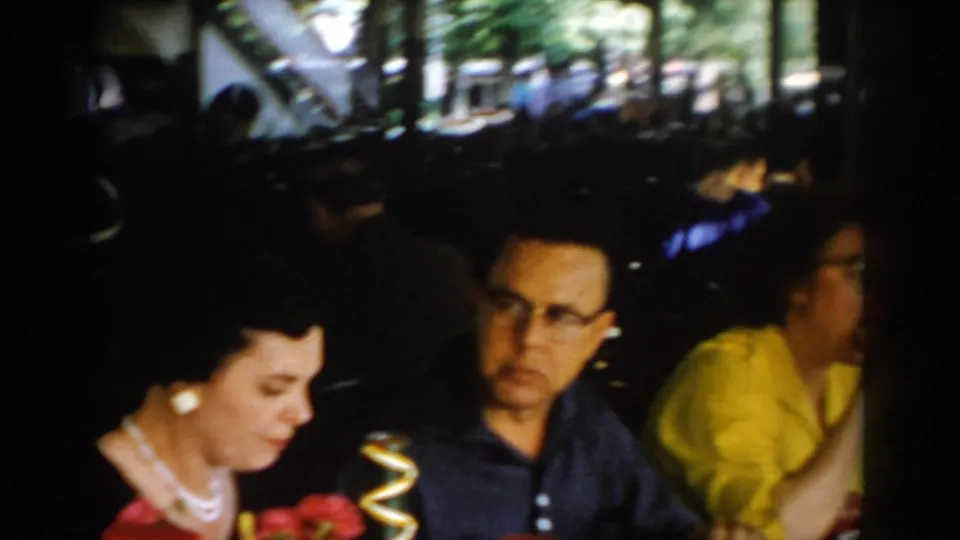 Adults dining in a covered patio restaurant, circa 1965 Saint Louis — woman in pearl necklace and red top, man in dark shirt with cat-eye glasses, woman in yellow blouse visible.