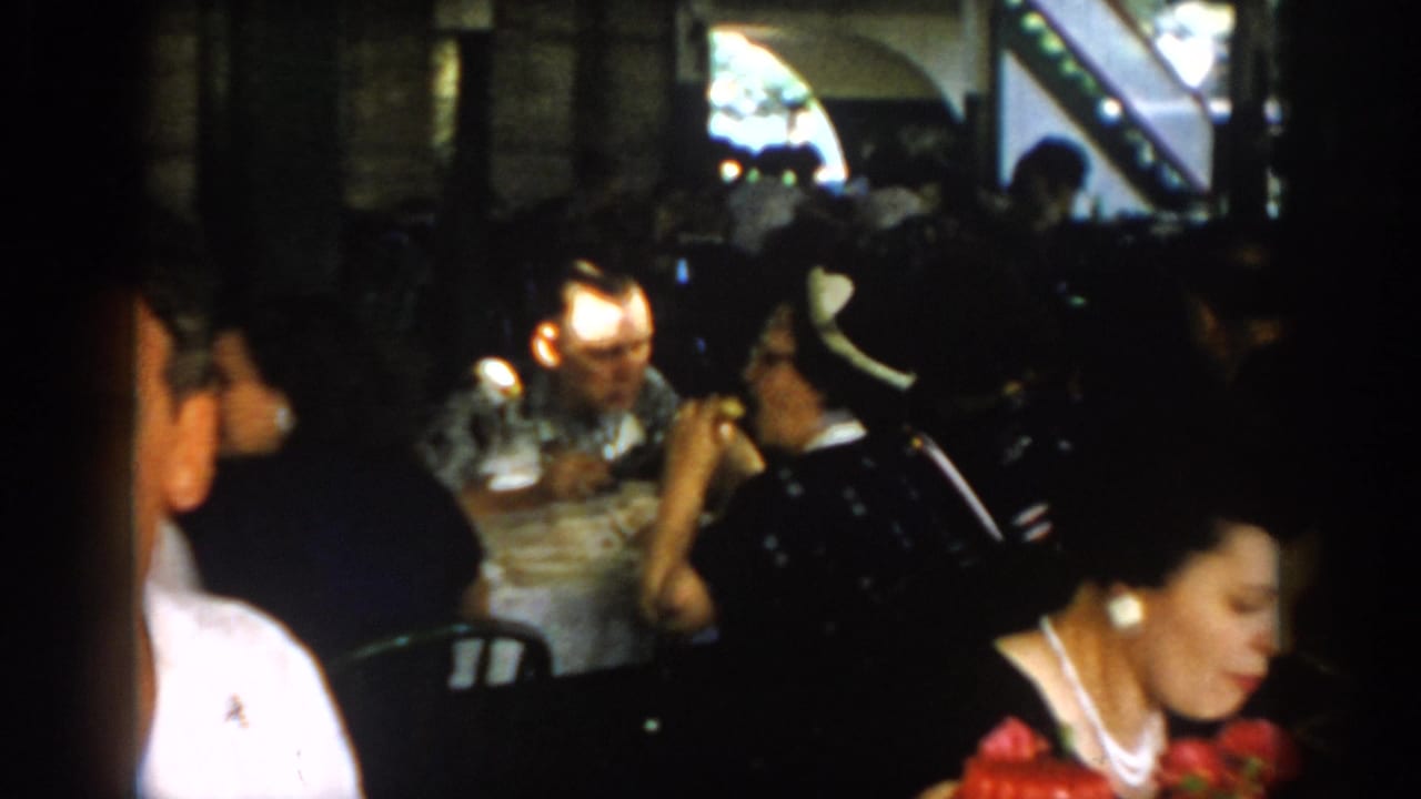 Candid view of adults dining in a covered patio, possibly Saint Louis, circa 1965 — woman with pearl necklace at left edge, diners silhouetted against bright background window.