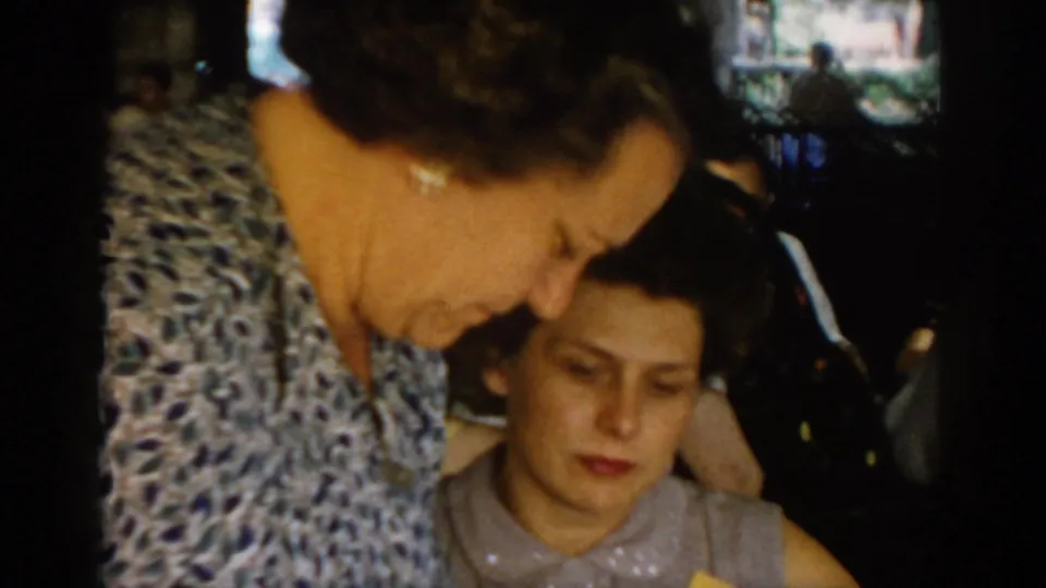 Two adult women at a restaurant table in a covered patio, circa 1965 Saint Louis — foreground figure in blue-grey patterned blouse, second woman leaning close beside her.