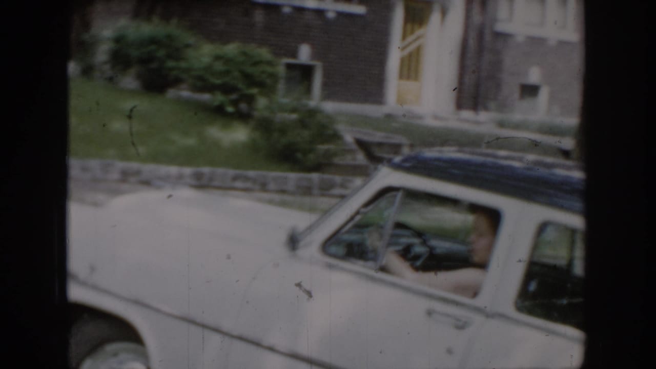 Woman driving a white 1960s-era American sedan with dark hardtop roof in a brick-house suburban setting, consistent with a mid-1960s St. Louis, Missouri home movie.