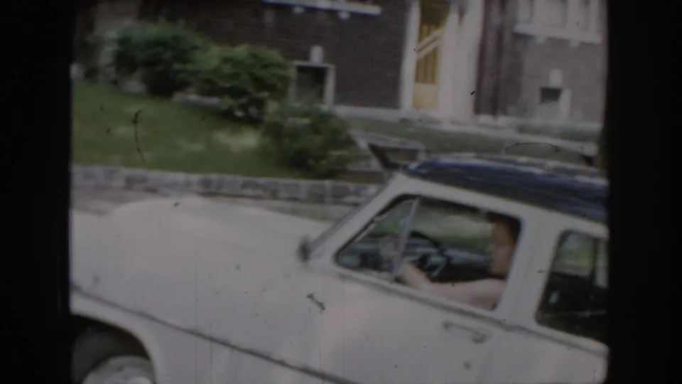 Woman driving a white 1960s-era American sedan with dark hardtop roof in a brick-house suburban setting, consistent with a mid-1960s St. Louis, Missouri home movie.