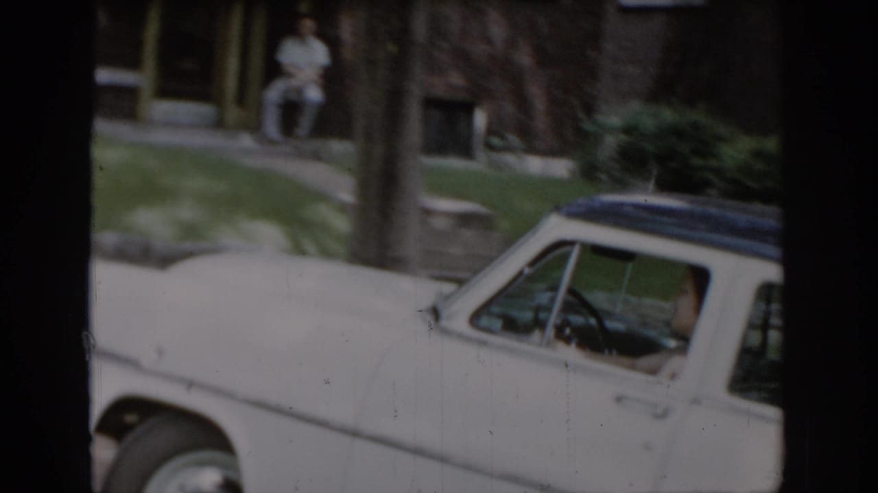 Wide view of a white 1960s American sedan in a leafy suburban setting with a man seated on porch steps in the background, consistent with St. Louis, Missouri, mid-1960s home movie footage.