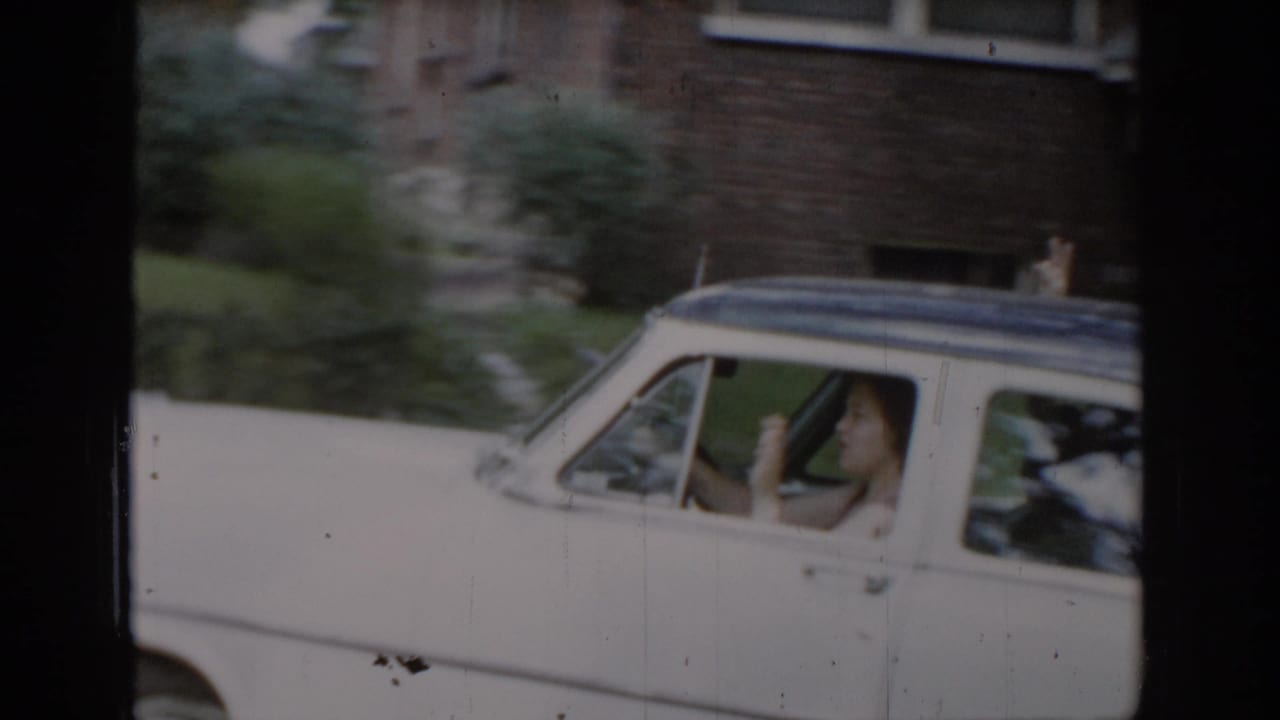 Woman waving from the open driver's window of a white 1960s American sedan passing a brick suburban house, consistent with St. Louis home movie footage, ca. 1965.