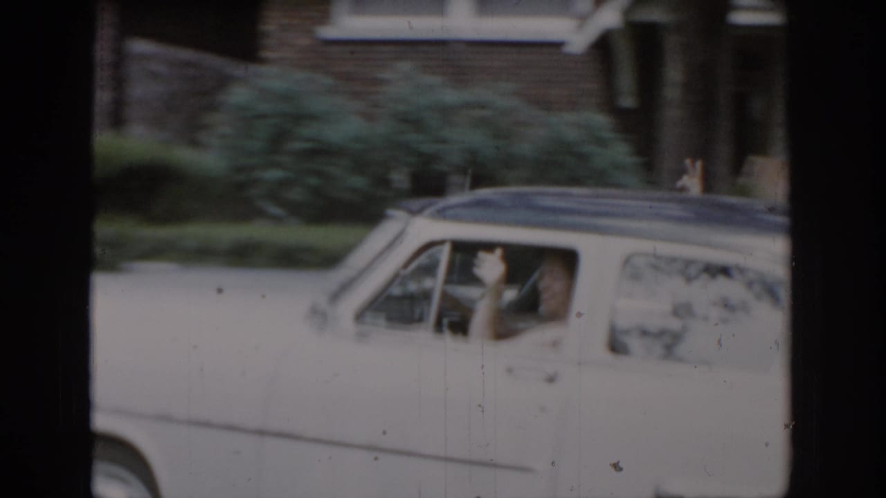 White 1960s American sedan pulling away, woman's arm extended in farewell wave through open window, brick house with decorative column visible, consistent with St. Louis suburban home movie, ca. 1965.