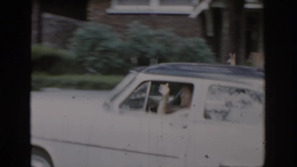 White 1960s American sedan pulling away, woman's arm extended in farewell wave through open window, brick house with decorative column visible, consistent with St. Louis suburban home movie, ca. 1965.