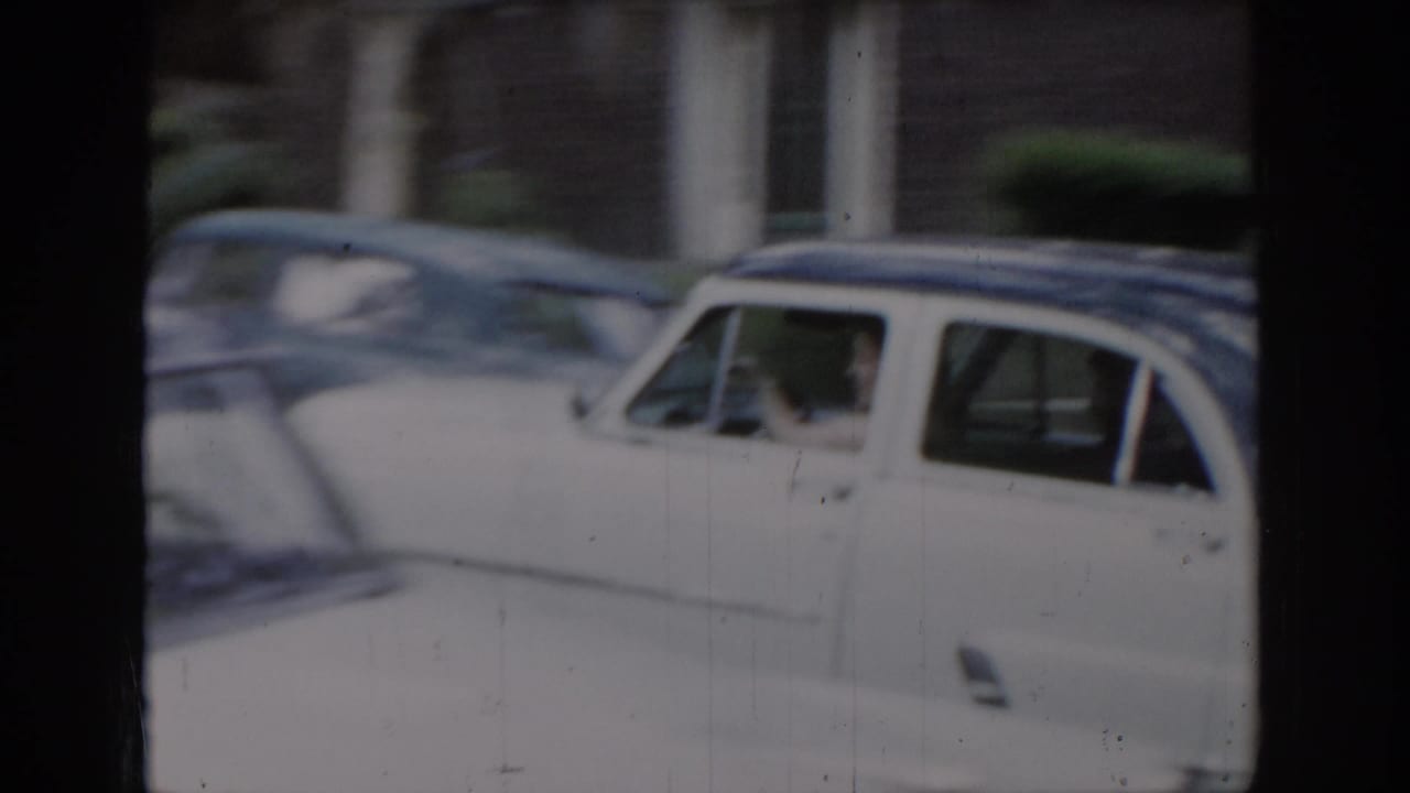 White classic American sedan parked in a residential driveway, with green foliage and brick building in background; 8mm film frame.