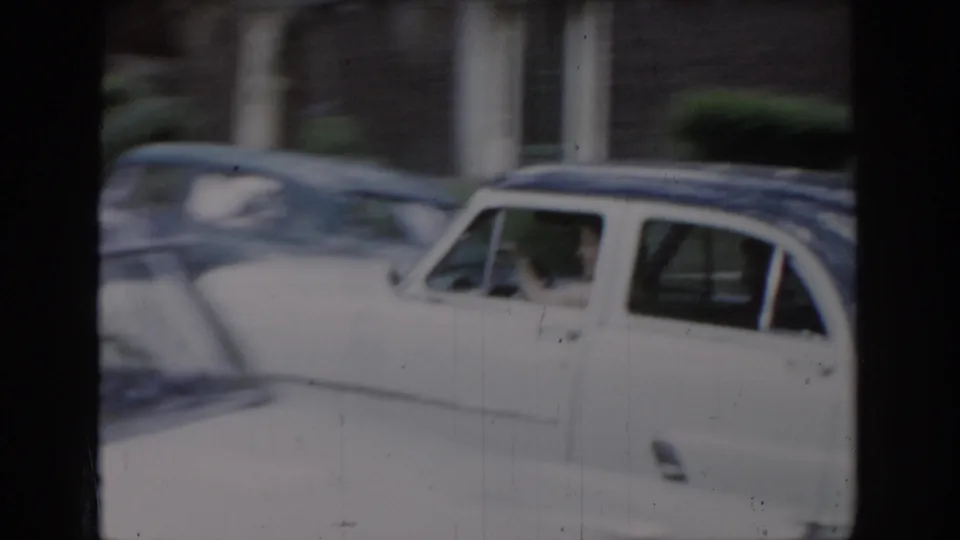 White late-1950s or early-1960s American sedan in a residential driveway with trees and brick building behind, from 8mm home movie footage attributed to Saint Louis, Missouri, c. 1965.