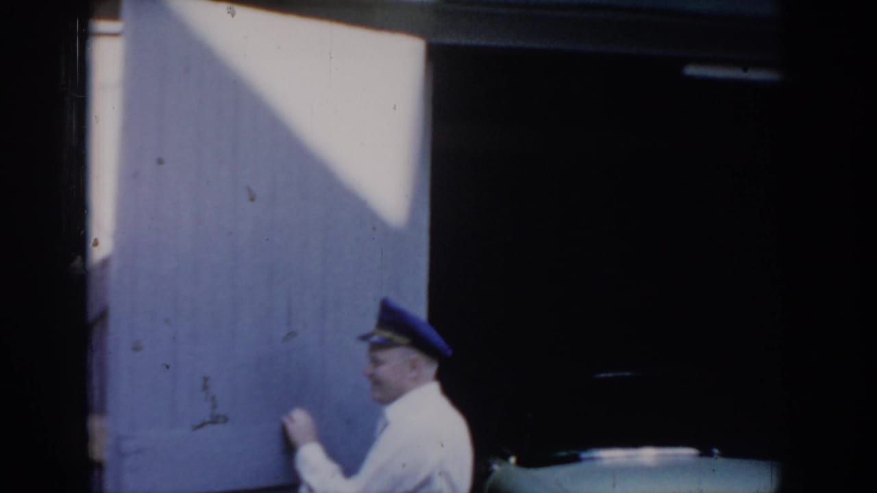 Man in white shirt and blue peaked cap opening a wooden barn-style garage door in bright sunlight, dark interior behind; 8mm home movie attributed to Saint Louis, Missouri, c. 1965.