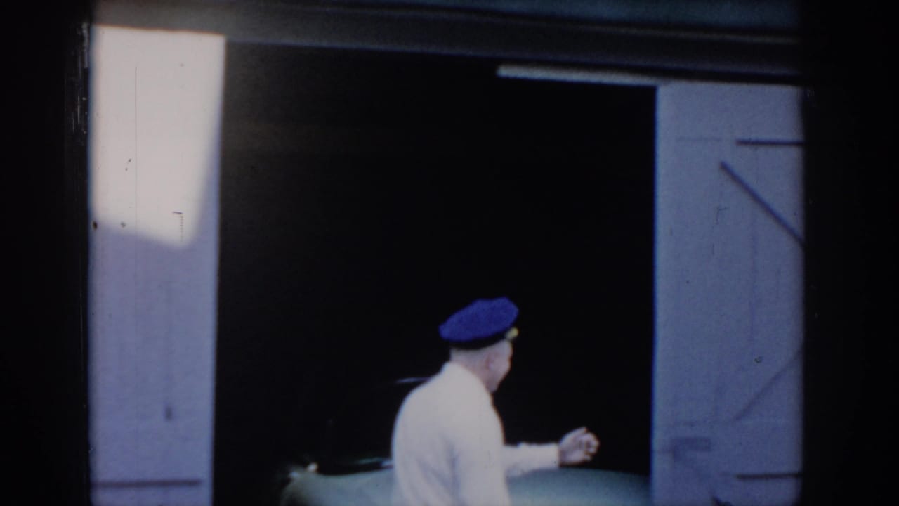 Man in white shirt and blue peaked cap pausing in an open barn garage doorway before entering, bright light from left; 8mm home movie attributed to Saint Louis, Missouri, c. 1965.