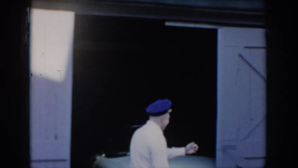 Man in white shirt and blue peaked cap pausing in an open barn garage doorway before entering, bright light from left; 8mm home movie attributed to Saint Louis, Missouri, c. 1965.