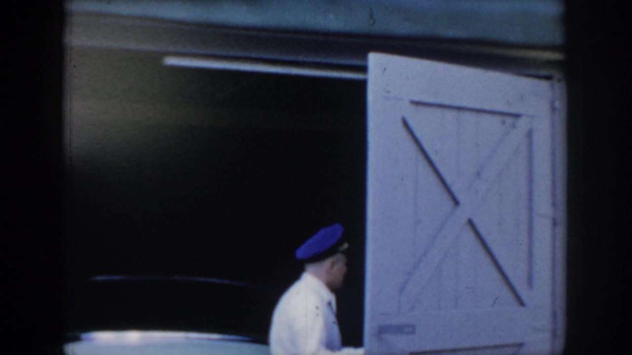 Man in white shirt and blue peaked cap entering a dark barn garage, white wooden barn doors with X-cross bracing at right; 8mm home movie attributed to Saint Louis, Missouri, c. 1965.