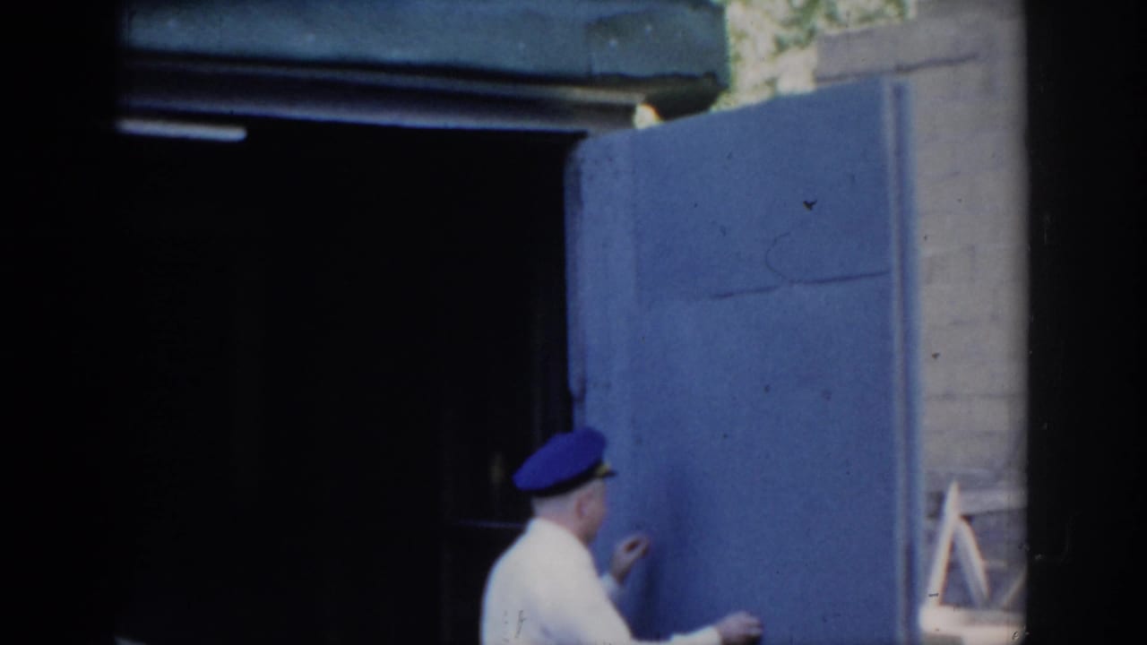 Man in white uniform and blue cap holding the open blue garage door; a wooden stepladder is visible at right.