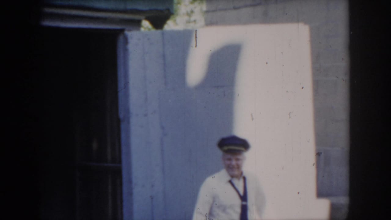 Uniformed man stepping into sunlight outside garage, tie and blue cap visible, cast shadow on cinder-block wall; Saint Louis, Missouri, circa 1965.