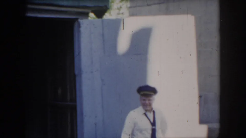 Uniformed man stepping into sunlight outside garage, tie and blue cap visible, cast shadow on cinder-block wall; Saint Louis, Missouri, circa 1965.