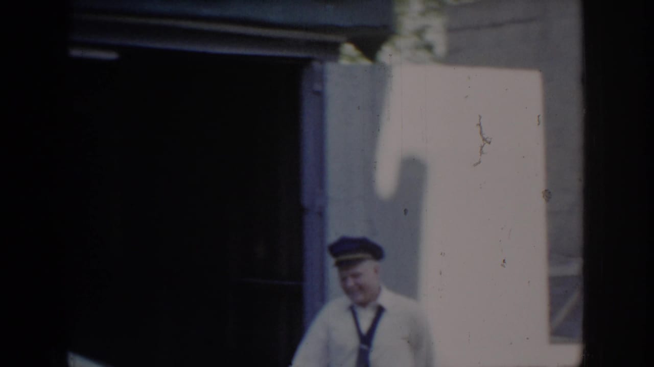 Smiling older man in white service uniform and blue peaked cap standing outside garage in bright sunlight; Saint Louis, Missouri, circa 1965, 8mm home movie.