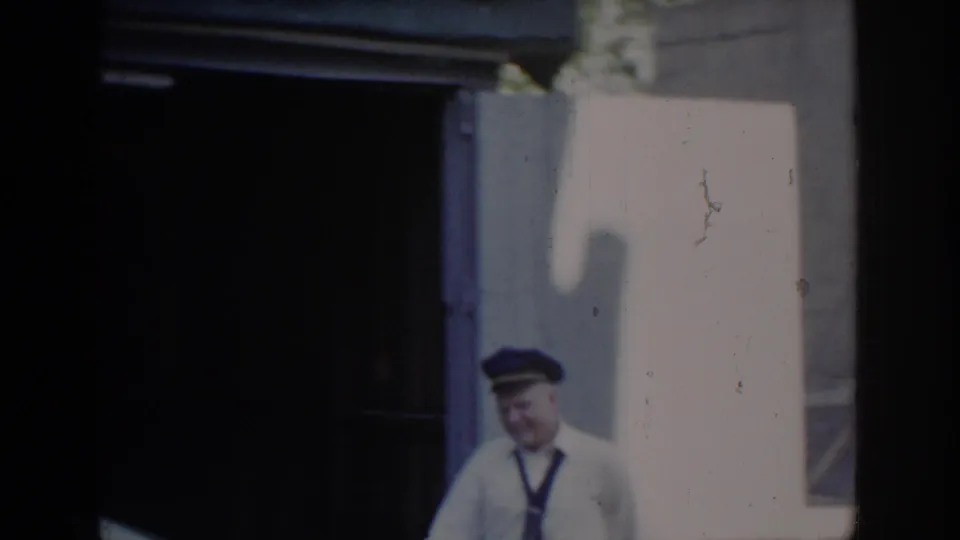 Smiling older man in white service uniform and blue peaked cap standing outside garage in bright sunlight; Saint Louis, Missouri, circa 1965, 8mm home movie.