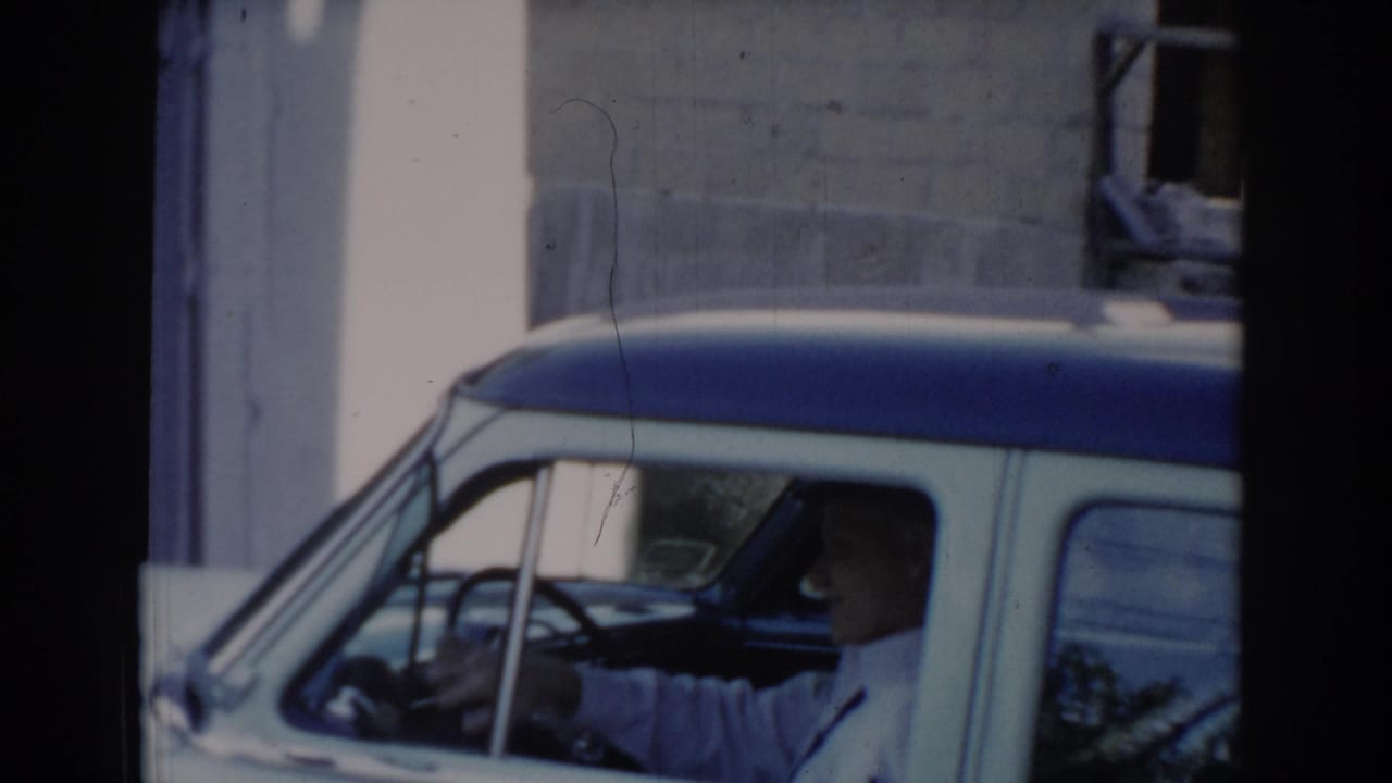 Front-side angle of two-tone blue and white sedan; man visible through driver's window, brick building in background.