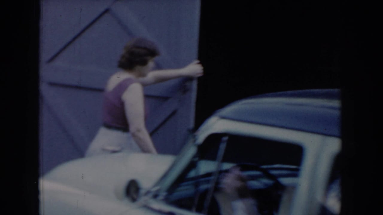 Woman opening a large wooden X-brace garage door beside a two-tone sedan in a residential driveway — circa 1965, Saint Louis, Missouri.