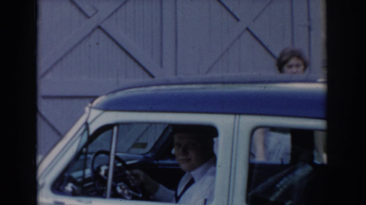 Man behind the wheel of a two-tone sedan framed through the windshield; wooden garage door and second figure in background — circa 1965, Saint Louis, Missouri.