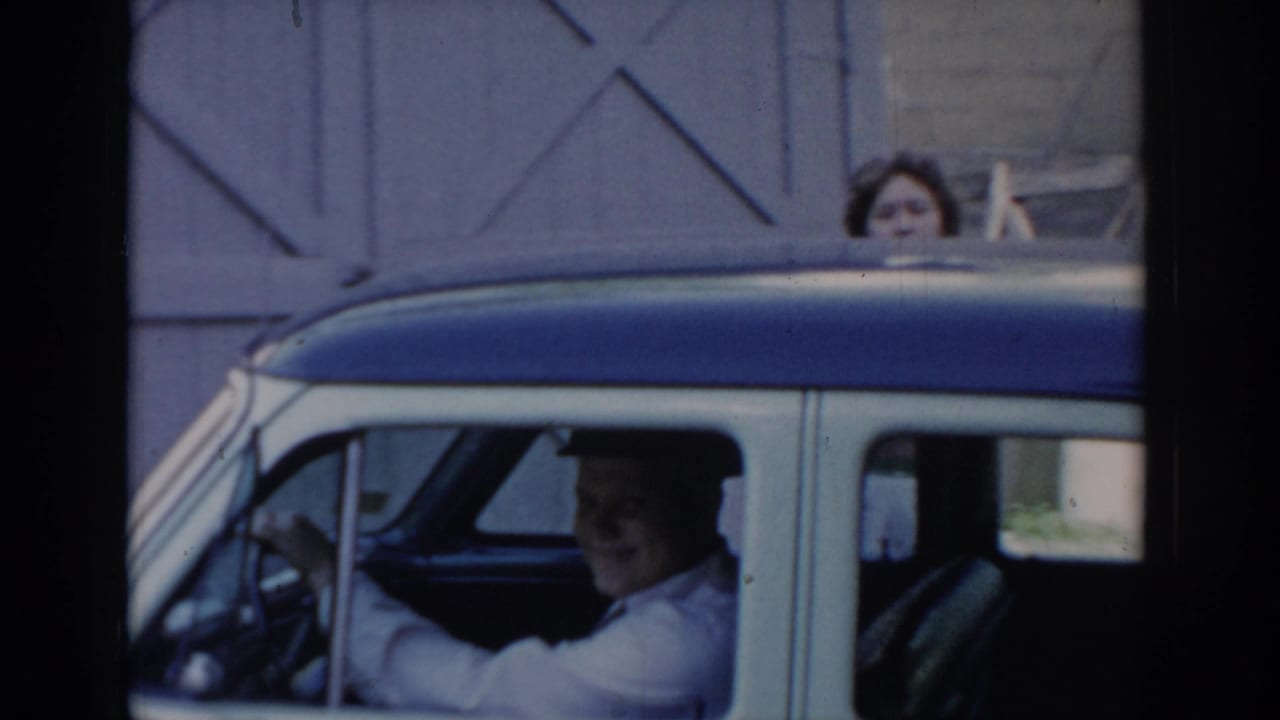 Man driving two-tone blue-and-white car with woman visible above roofline; cross-braced wooden barn door in background; 1960s home movie.