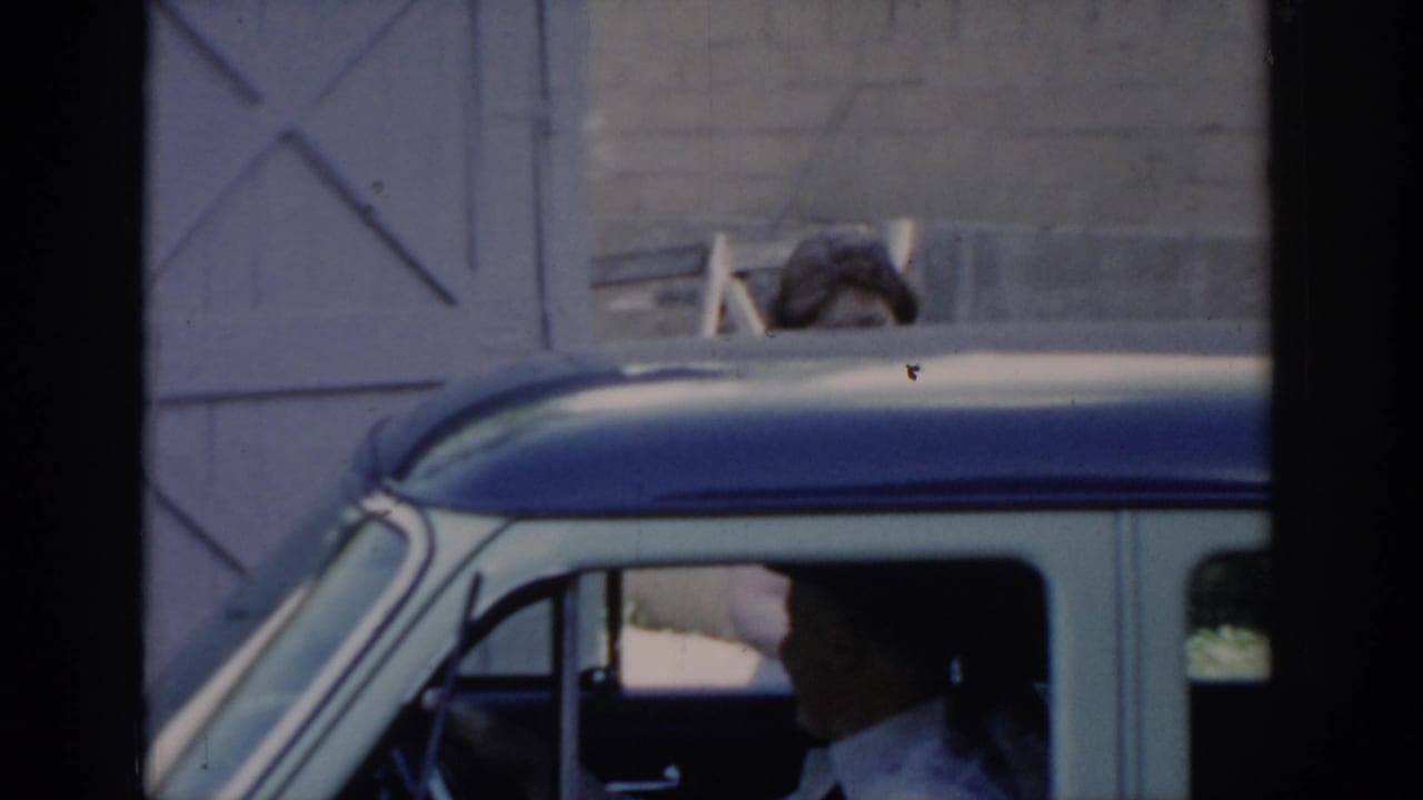 Man in white shirt visible through driver's window of two-tone 1960s car; figure above roofline against cross-braced barn door background.