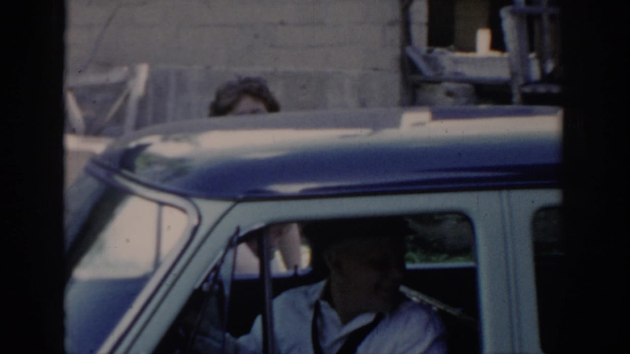 Woman with dark curly hair stands near a two-tone car; metal containers and cross-braced barn door visible in background; consistent with 1960s home movie setting.