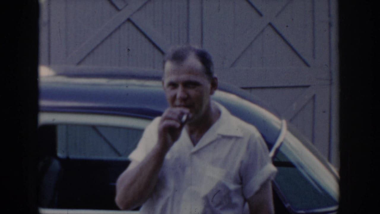 Man in white short-sleeve collared shirt smoking near a two-tone car; cross-braced barn doors behind; candid 1960s home movie moment.