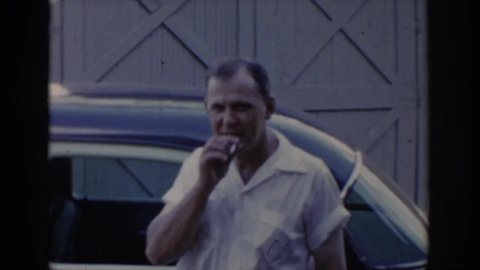 Man in white short-sleeve collared shirt smoking near a two-tone car; cross-braced barn doors behind; candid 1960s home movie moment.